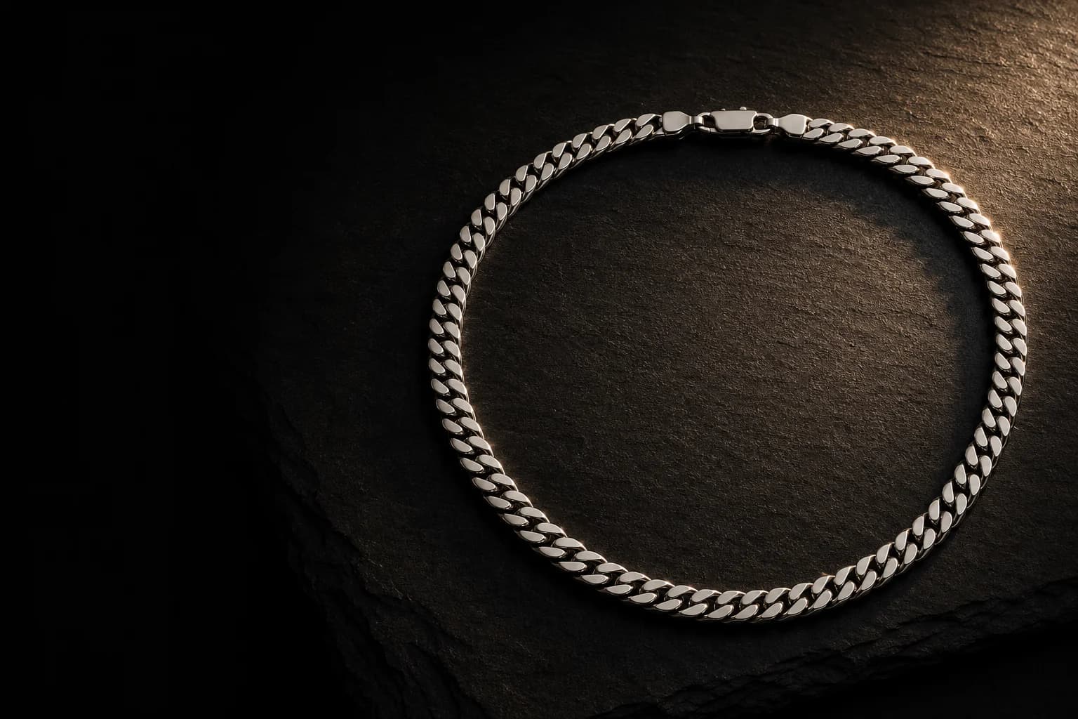A polished steel Cuban link chain coiled on black slate, caught by a single warm raking light