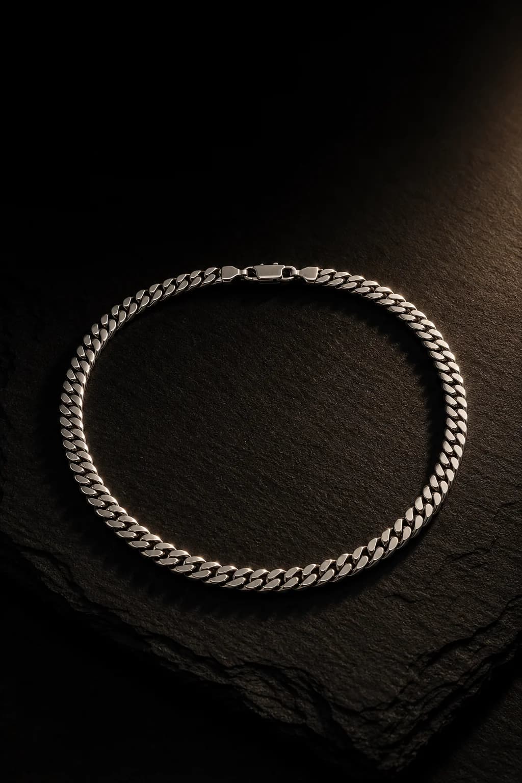 A polished steel Cuban link chain coiled on black slate, caught by a single warm raking light