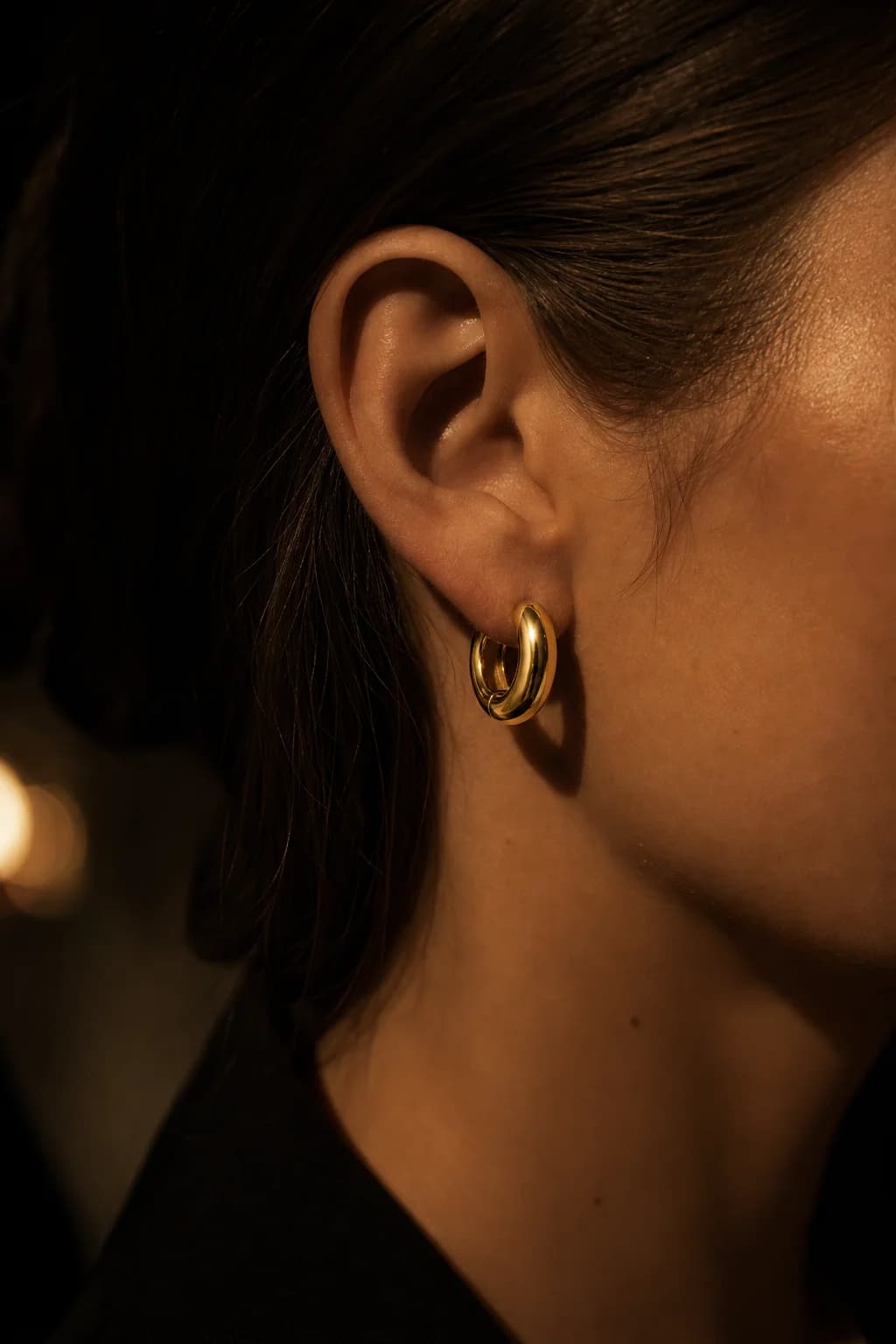 A chunky gold huggie hoop worn on an ear, hair tucked back, in warm low light